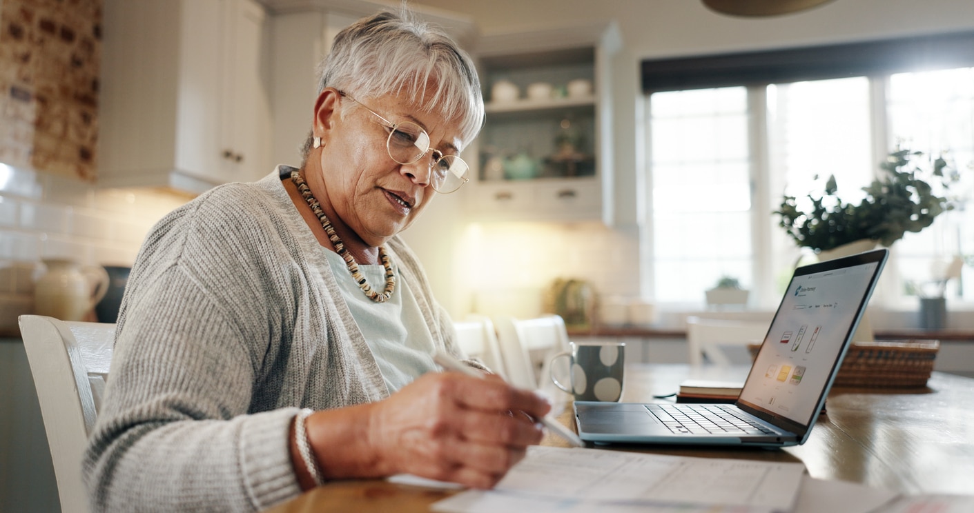 A senior woman sits at a table, surrounded by paperwork, a laptop open in front of her. She is reviewing financial documents—likely inquiring about fatca affecting foreign pensions. The setting suggests home, and the mood is one of careful planning and concern for financial details.